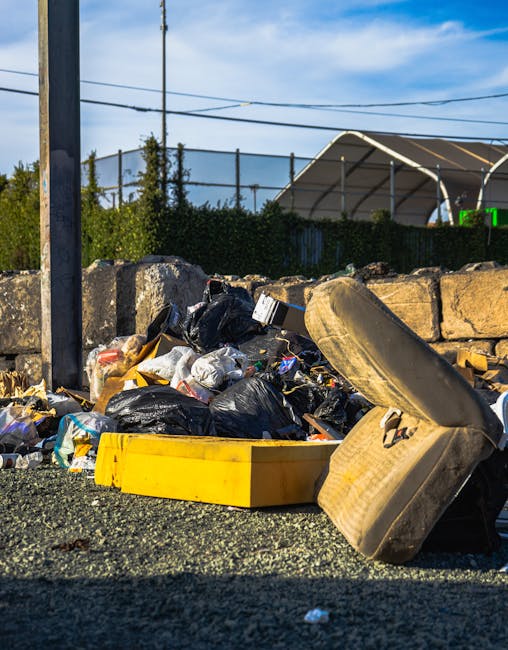 A collection of mixed rubbish and waste materials accumulated on the side of a road or driveway, including black and white plastic trash bags, a large yellow plastic bin, a discarded old car tire, and various unlabelled packaging and debris. The rubbish is piled in front of a rough stone wall, with some of the waste spilling onto a gravel surface. Behind the waste, a chain-link fence runs parallel to the scene, with green shrubbery and trees visible beyond the fence. To the right, part of a large, industrial-style tent or canopy with a metal frame and fabric covering can be seen, possibly part of a waste collection or storage area. Overhead, a clear blue sky with a few wispy clouds and visible utility wires suggest a bright, sunny day. The scene reflects a typical example of waste accumulation requiring rubbish removal or clearance, with the materials suitable for outsourced collection services such as House Clearance Battersea, highlighting the importance of proper waste disposal.