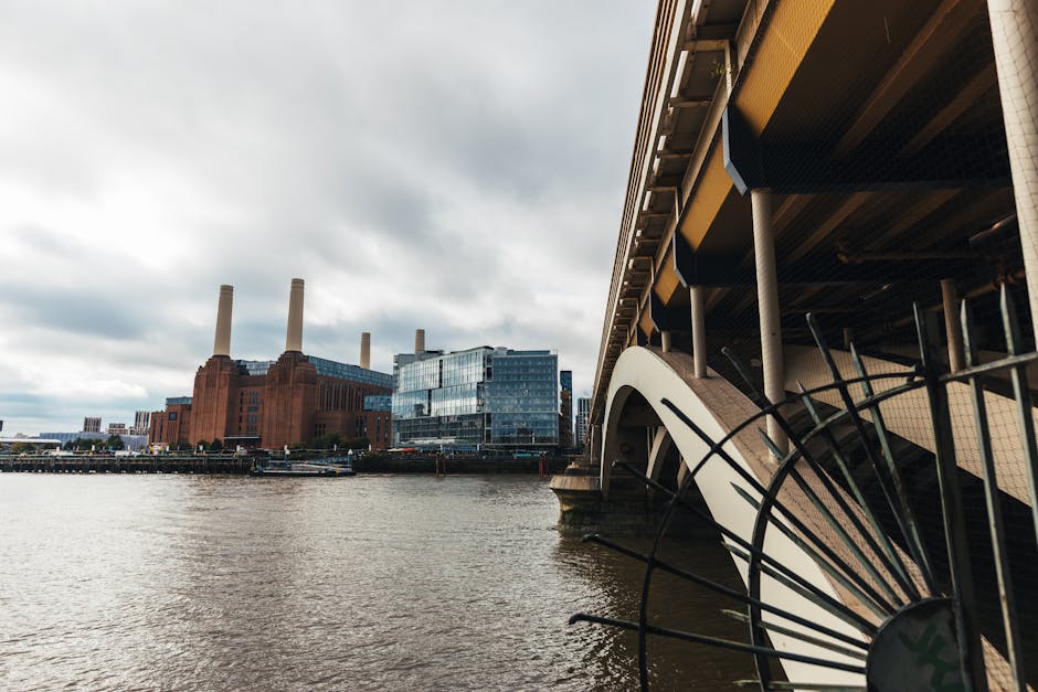 A large historic building situated along the riverbank, constructed from reddish-brown brick with a textured surface, featuring tall, cylindrical chimneys rising above the structure and flanked by modern glass-fronted buildings. The building's facade displays vertical window arrangements and architectural detailing typical of industrial or power station architecture. In the foreground, a section of the river with gentle ripples and a small boat or barge floating near the riverbank is visible, with the water reflecting the overcast sky. Behind the building, the sky appears cloudy and gray, contributing a muted lighting atmosphere. The scene suggests an urban environment where historic infrastructure combines with contemporary development, indirectly highlighting the importance of responsible waste management and the potential for independent rubbish removal or site clearance to maintain such areas. The presence of House Clearance Battersea in a relevant context of private waste handling emphasizes the importance of professional rubbish services for urban preservation and efficient disposal.