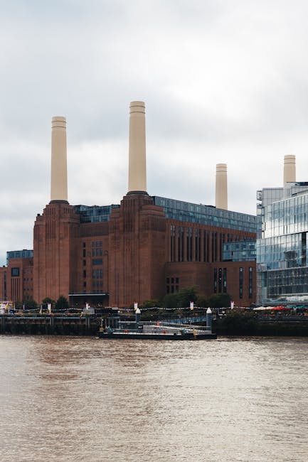 A large historic power station with a prominent reddish-brown brick facade, featuring vertical windows and four tall, cream-colored chimney stacks extending upward, situated adjacent to a river with a small boat moored along the shoreline. To the right, there are modern glass-fronted office buildings reflecting the overcast sky, which is filled with grey clouds. The scene captures an urban riverside environment, where the power station stands as a notable industrial silhouette against the water, and the nearby contemporary structures suggest a mixed-use cityscape. In the context of rubbish removal or waste management services, the image highlights the potential for on-site clearance or private collection in an industrial or urban setting, consistent with alternative waste handling options outside municipal services. The surroundings appear calm, with no active construction or loading activity visible, emphasizing the static nature of the scene.