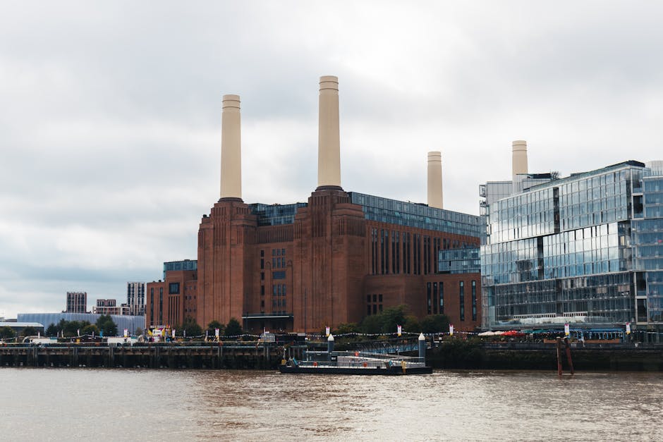 A large industrial building with a reddish-brown brick exterior situated along a waterfront, featuring tall, cream-colored chimneys extending upwards from the roof. The building appears robust with a textured surface and multiple vertical windows arranged in a grid pattern. To the right, there is a modern glass-fronted structure with reflective panels that mirror the cloudy sky, creating a contrast in architectural style. The foreground shows a section of water, likely a river or canal, with a small dock or quay area where a road or pathway runs parallel to the water’s edge. Alongside the shoreline, there are barriers or bollards for safety, and some trees or shrubbery are visible beyond, providing minimal greenery. The overall atmosphere is overcast with diffused light, emphasizing the industrial and urban setting, which is relevant to themes of waste management or alternative disposal methods that companies like House Clearance Battersea may facilitate through their rubbish removal services.