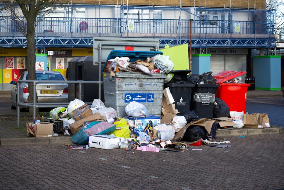 A collection of overflowing rubbish bins and scattered waste on a paved sidewalk in front of a commercial building, including large black and red skip bags filled with mixed waste, open cardboard boxes, and various discarded packaging materials. The scene features a mix of crumpled paper, plastic bags, and discarded objects spilling onto the ground, with a small grey car parked nearby and a low metal railing separating the waste area from the roadway. In the background, the building facade has storefronts with signs and a blue construction scaffold covering part of the upper levels, suggesting ongoing maintenance or renovation work. The environment appears urban, with natural daylight illuminating the scene, highlighting the cluttered and untidy state of the waste area, which House Clearance Battersea may handle as part of private rubbish removal or on-site clearance services in the Battersea area.