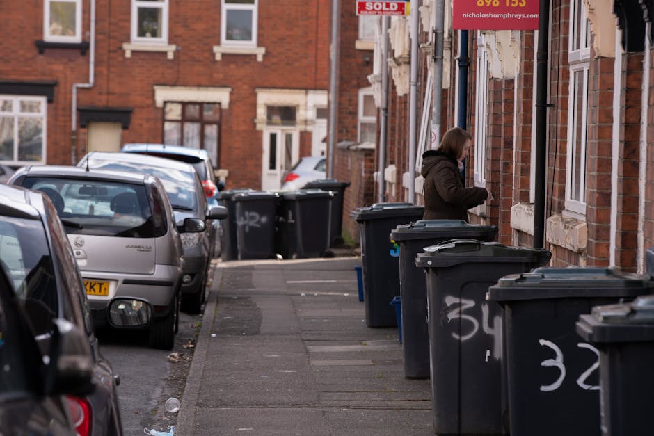 A narrow urban street scene featuring a row of dark grey wheelie bins lined up along the pavement outside red brick residential buildings. The bins are positioned close to the building facade, with some stacked slightly behind others, and have white spray-painted numbers on their fronts. A woman wearing a dark brown coat stands near one bin, appearing to be handling or accessing it. Several parked cars in various shades of grey and silver occupy the street, with their rear or front ends facing the viewer. The background shows more brick houses with white-framed windows and a signboard hanging from one building. The environment appears to be during daylight hours with natural lighting illuminating the scene, and residual litter can be seen on the pavement. The setting suggests a typical residential street where private waste collection services like house clearance companies might be involved, reflecting a context of independent rubbish disposal outside of council services. House Clearance Battersea's expertise in rubbish management is subtly supported by the orderly arrangement and access to these waste containers, often used for on-site clearance or private waste handling across Battersea.