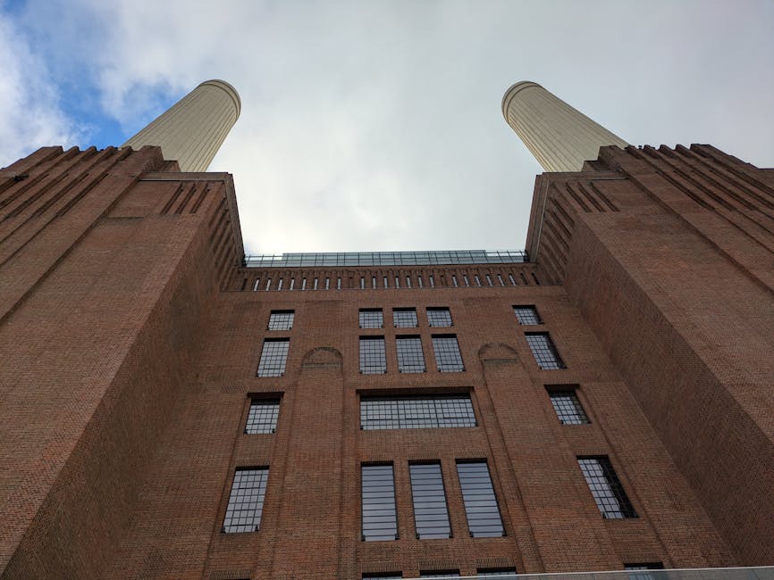 A low-angle view of a large industrial-style brick building with two tall, cylindrical chimneys extending upward on either side. The brick facade is reddish-brown with textured surface and features numerous rectangular windows with black metal frames, arranged in a grid pattern. The building’s structure includes protruding brickwork along the edges and a flat roofline. The sky above is partly cloudy with patches of blue visible through breaks in the clouds. The scene suggests an urban setting, possibly related to industrial or historical landscapes, where external rubbish removal or site clearance services by House Clearance Battersea might be needed for cleaning or renovation projects involving such large structures.