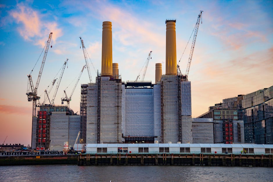 An image of Battersea Power Station in London during sunset, with a clear sky painted in soft pink and blue hues. The historic building features large, symmetrical chimneys with a beige, slightly weathered finish and a metallic sheen at the top. The power station's facade is constructed from red brick and grey concrete, with sections wrapped in protective scaffolding and white sheeting indicating ongoing maintenance or restoration work. Surrounding the power station are several construction cranes with long, lattice arms extending into the sky, positioned on the left and right sides of the structure, actively involved in ongoing development. The station is situated on a body of water, with a wooden pier or dock visible at the water’s edge, supporting the building. The overall scene captures the contrast between industrial architecture and the natural beauty of the sunset, reflecting the ongoing urban redevelopment in the Battersea area. House Clearance Battersea may offer alternative waste handling services for construction or renovation debris related to projects at such sites, aligning with independent rubbish removal options.