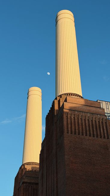 A low-angle view of a large industrial-style brick building with two tall, cylindrical chimneys extending upward on either side. The brick facade is reddish-brown with textured surface and features numerous rectangular windows with black metal frames, arranged in a grid pattern. The building’s structure includes protruding brickwork along the edges and a flat roofline. The sky above is partly cloudy with patches of blue visible through breaks in the clouds. The scene suggests an urban setting, possibly related to industrial or historical landscapes, where external rubbish removal or site clearance services by House Clearance Battersea might be needed for cleaning or renovation projects involving such large structures.