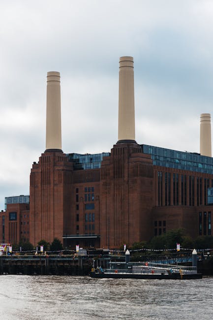 A large industrial building, likely a former power station, constructed from reddish-brown brick with a stepped, Art Deco style façade. The structure features three tall, cylindrical chimneys made of a similar material, rising prominently above the building's roofline. The building's exterior includes numerous rectangular windows, some of which are tinted or reflective, and sections with vertical and horizontal detailing in the brickwork. In the foreground, there's a body of water reflecting the building's lower portion, with a small boat or barge docked along the water’s edge. The scene is set outdoors under a cloudy sky, with a few trees visible at the base of the building, and a minimal amount of surrounding infrastructure such as fencing or railings. The overall scene underscores an industrial environment, and the image could relate to the context of managing or removing waste or debris from old industrial sites, aligning with private or alternative waste removal methods that occur on-site or nearby. The photo exemplifies the type of environment where rubbish removal services might be employed during building clearance or site clean-up processes, such as handled by companies like House Clearance Battersea.