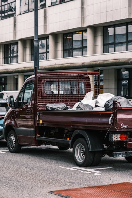 A burgundy flatbed truck parked on an urban street in front of a modern building with large glass windows and concrete panels. The truck's cargo bed is filled with various types of waste, including black plastic rubbish bags, some white and gray, that appear loosely packed and slightly crumpled. The waste material is stacked and piled, with some bags leaning against a metal mesh safety barrier at the back of the truck bed. The vehicle is situated near a street lamp, and other parked cars are visible in the background, indicating a city environment. The scene is captured during daylight with natural lighting, reflecting typical outdoor conditions. This image relates to independent waste collection and rubbish removal, a service that complements or provides alternatives to standard municipal disposal options, often used during house or estate clearance in residential or commercial areas, such as Battersea, near Clapham Junction.