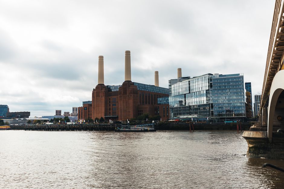 A large industrial building, likely a former power station, constructed from reddish-brown brick with a stepped, Art Deco style façade. The structure features three tall, cylindrical chimneys made of a similar material, rising prominently above the building's roofline. The building's exterior includes numerous rectangular windows, some of which are tinted or reflective, and sections with vertical and horizontal detailing in the brickwork. In the foreground, there's a body of water reflecting the building's lower portion, with a small boat or barge docked along the water’s edge. The scene is set outdoors under a cloudy sky, with a few trees visible at the base of the building, and a minimal amount of surrounding infrastructure such as fencing or railings. The overall scene underscores an industrial environment, and the image could relate to the context of managing or removing waste or debris from old industrial sites, aligning with private or alternative waste removal methods that occur on-site or nearby. The photo exemplifies the type of environment where rubbish removal services might be employed during building clearance or site clean-up processes, such as handled by companies like House Clearance Battersea.